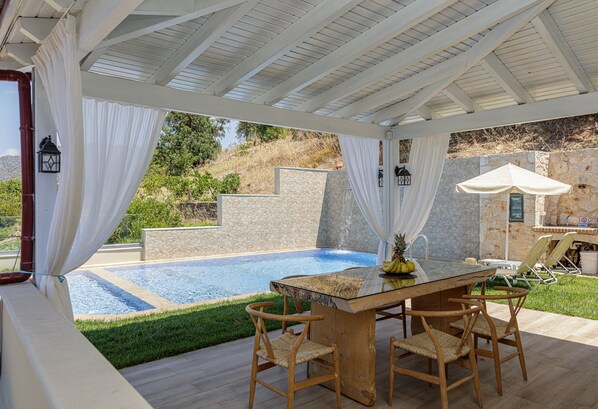 Zona de piscina al aire libre con una mesa y sillas de vidrio, rodeada de cortinas blancas y muros de piedra.