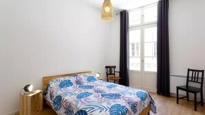 Una camera da letto con un letto, un comodino, una sedia, una lampada a sospensione e una finestra con tende.