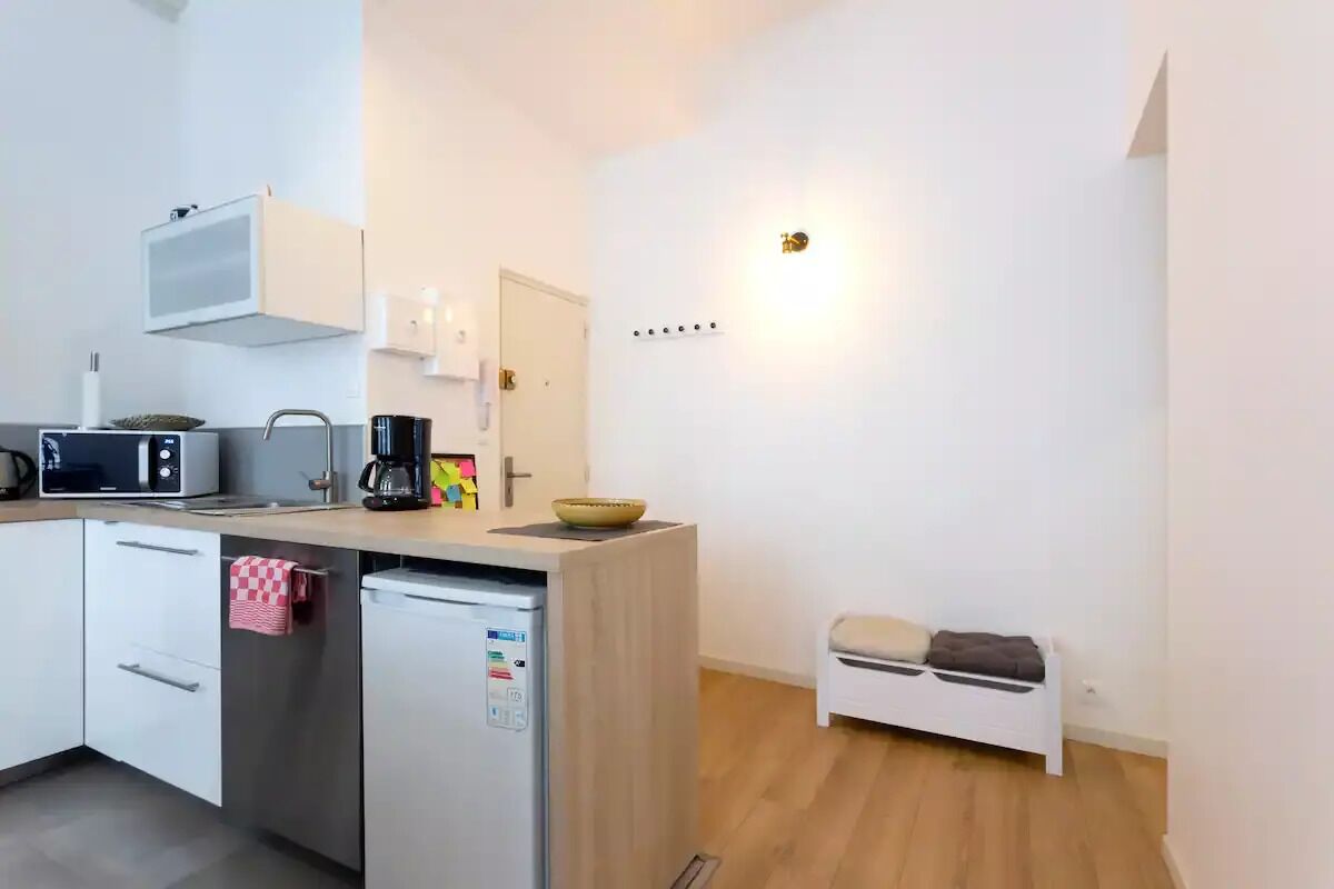 A compact kitchen with a microwave, coffee maker, and a small sink. There is a white cabinet with a towel hanging on it, and a small stool near the sink.