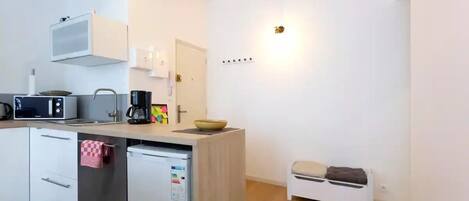 A compact kitchen with a microwave, coffee maker, and a small sink. There is a white cabinet with a towel hanging on it, and a small stool near the sink.