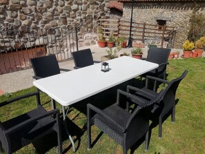 A rectangular outdoor table with a white top and black wicker chairs, surrounded by potted plants and a stone wall in the background.
