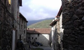 A narrow stone alley with traditional stone buildings, a parked car, and a view of a hill in the distance.