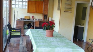 Un patio cubierto con una mesa dispuesta para comer, rodeado de sillas de mimbre. Hay una maceta con flores rojas sobre la mesa.