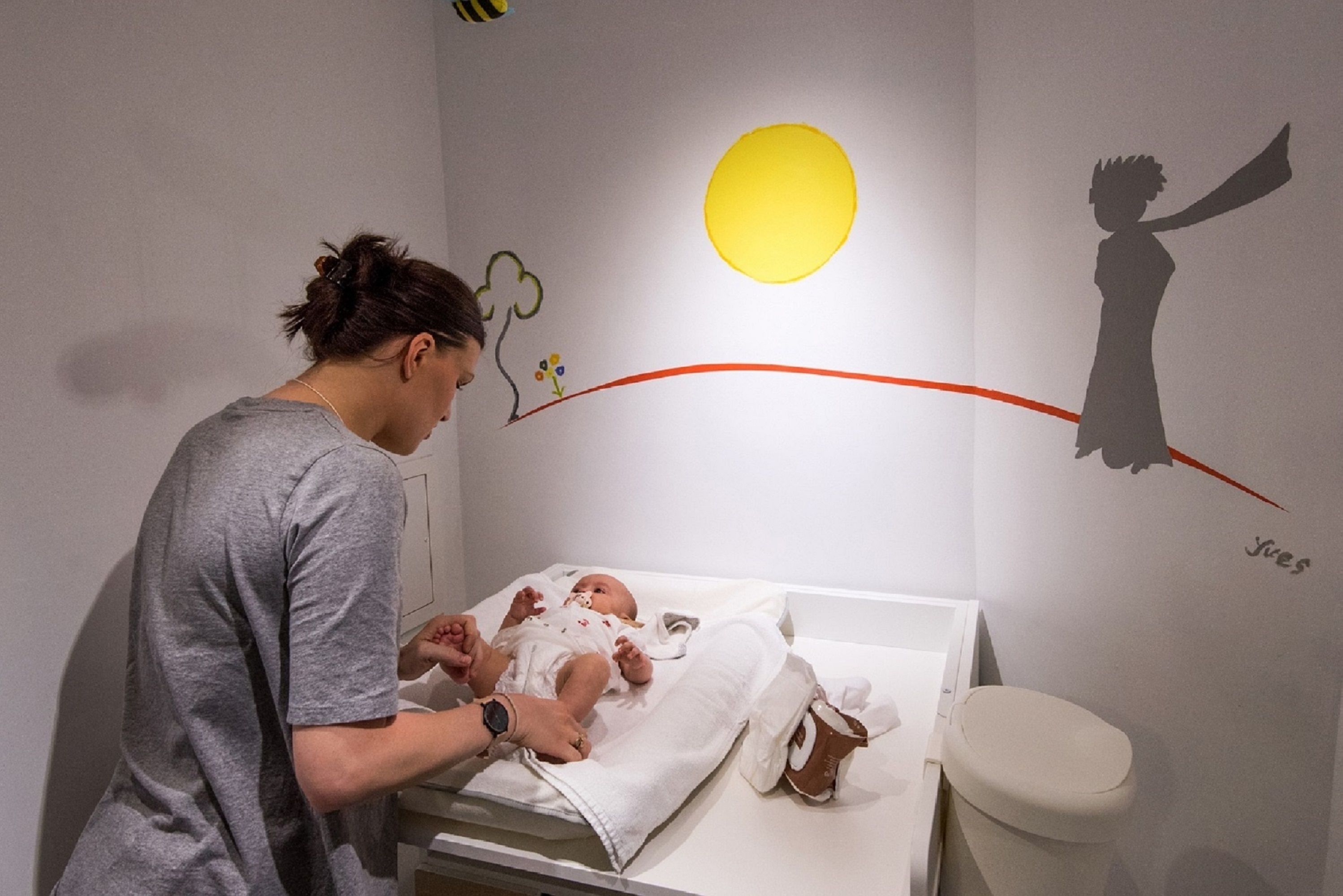 A room with a crib, a baby, and a wall with a sun and a silhouette of a person.