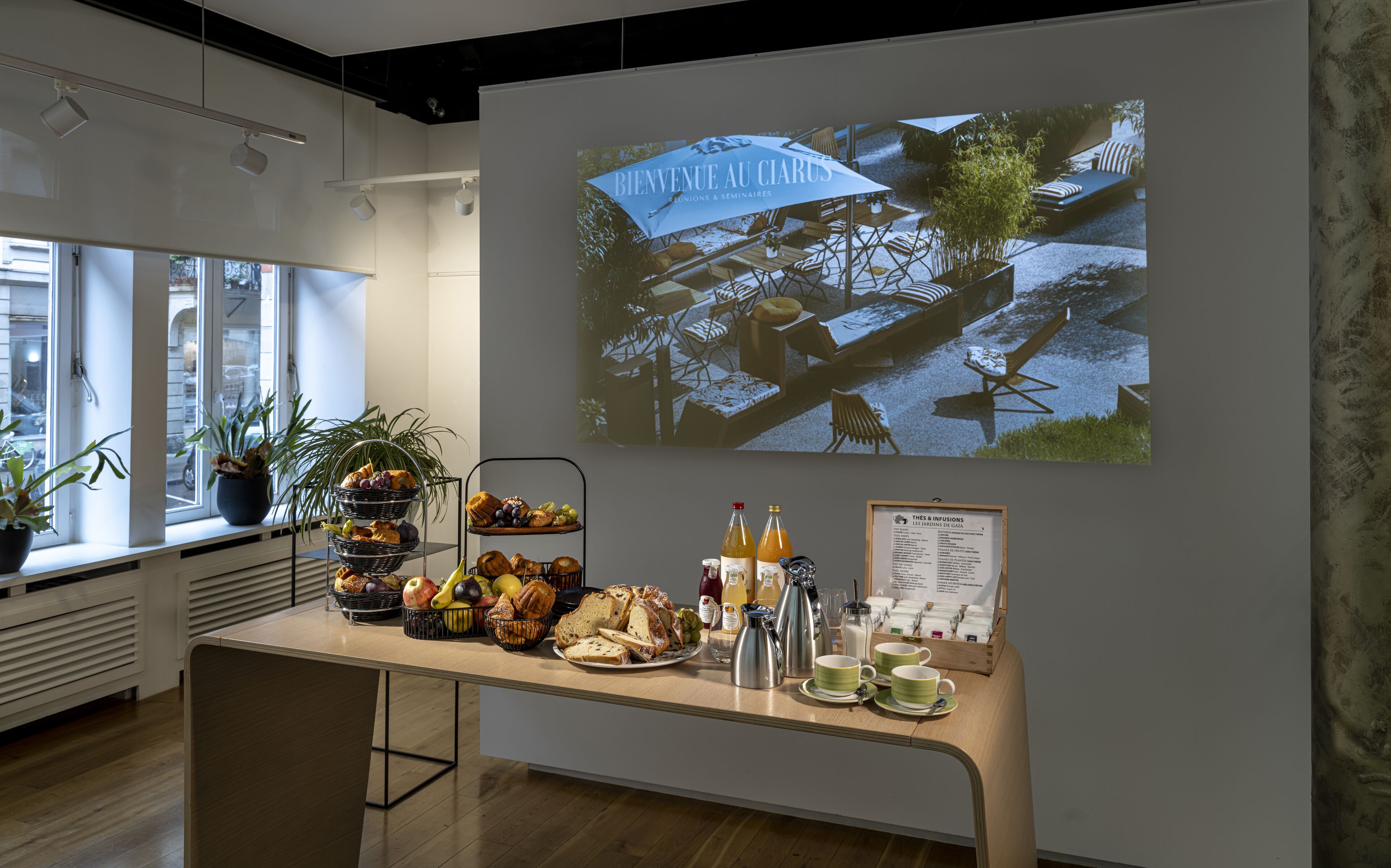 Zona de recepción con una mesa de buffet, una pantalla de proyección mostrando un mensaje de bienvenida y una ventana con persianas.