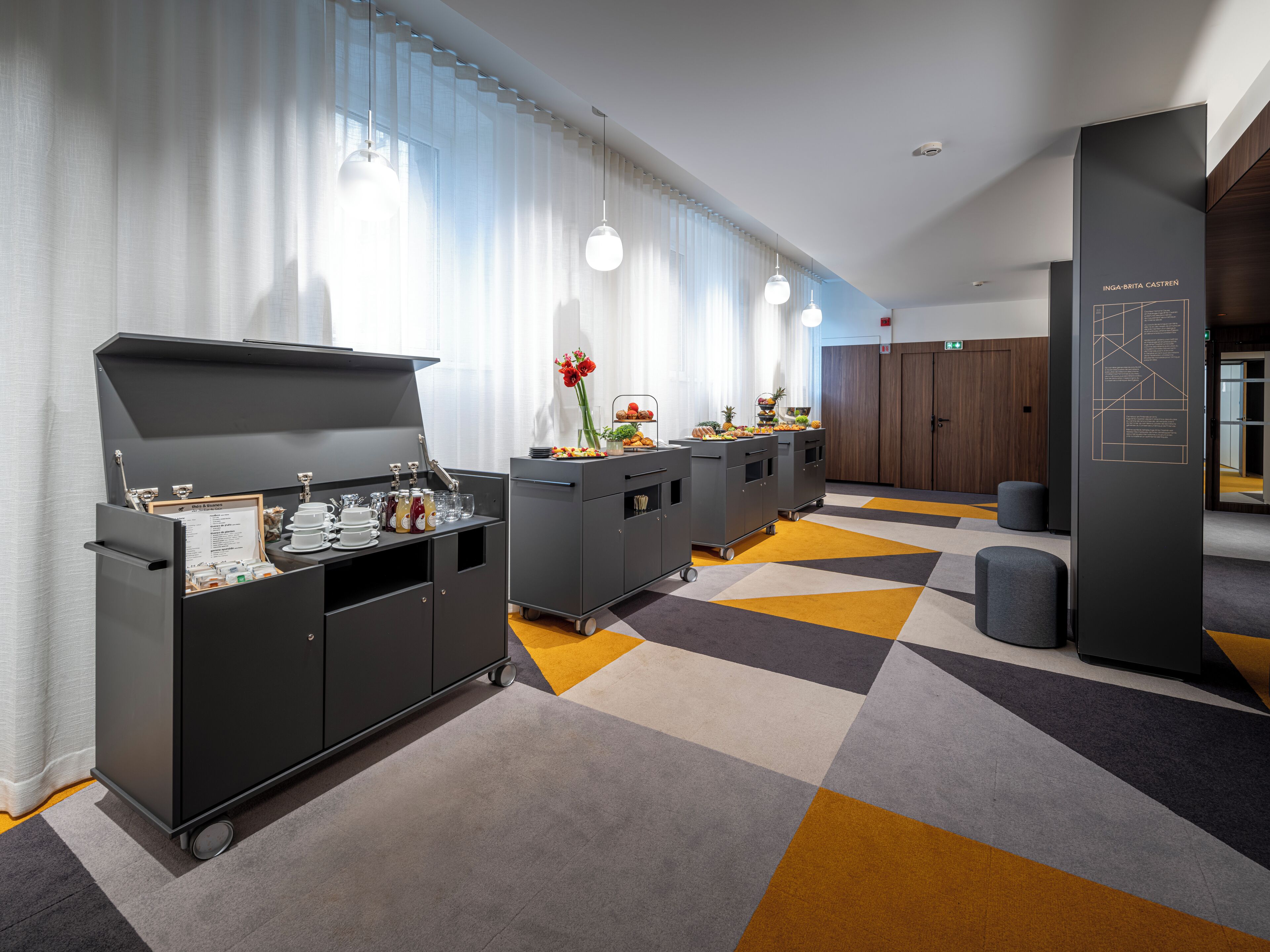 A modern hotel lobby with a kitchenette area, including a grill and beverage dispensers, and a seating area with a patterned carpet.