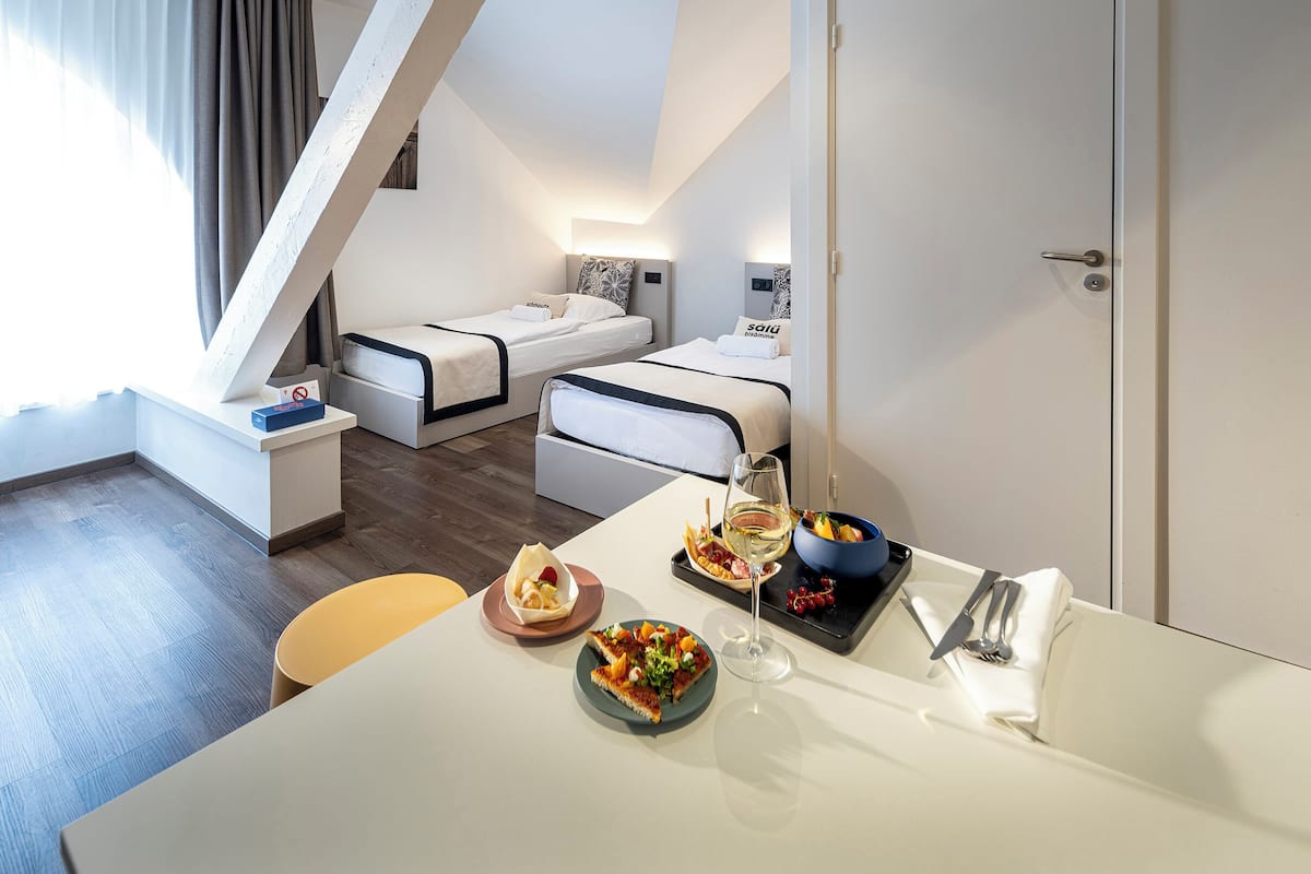A modern hotel room with a lofted sleeping area, a dining table with food and drink, and a large window with curtains.