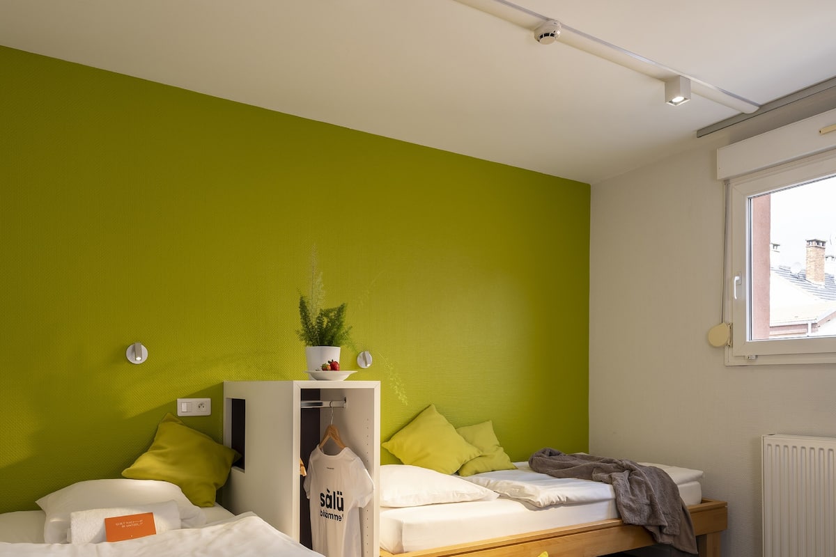 A modern hotel room with a green accent wall, a wooden bed frame, a bedside table, and a window with a view of the outside.