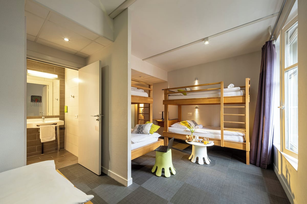 A hotel room with a bunk bed, a desk with a laptop, a small stool, and a bathroom with a mirror and sink.