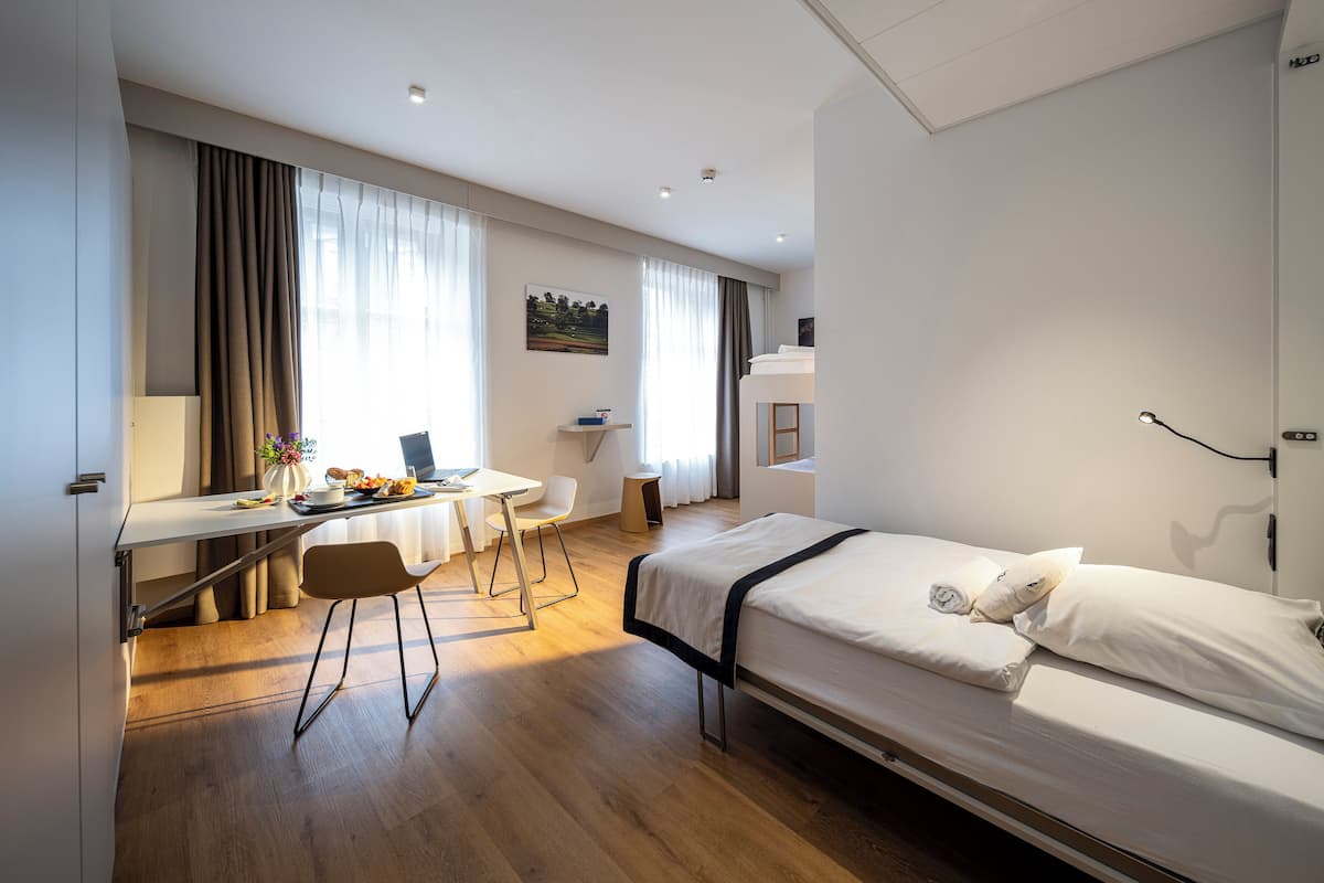 A modern hotel room with a bed, desk, chair, and a small table with a laptop and food.