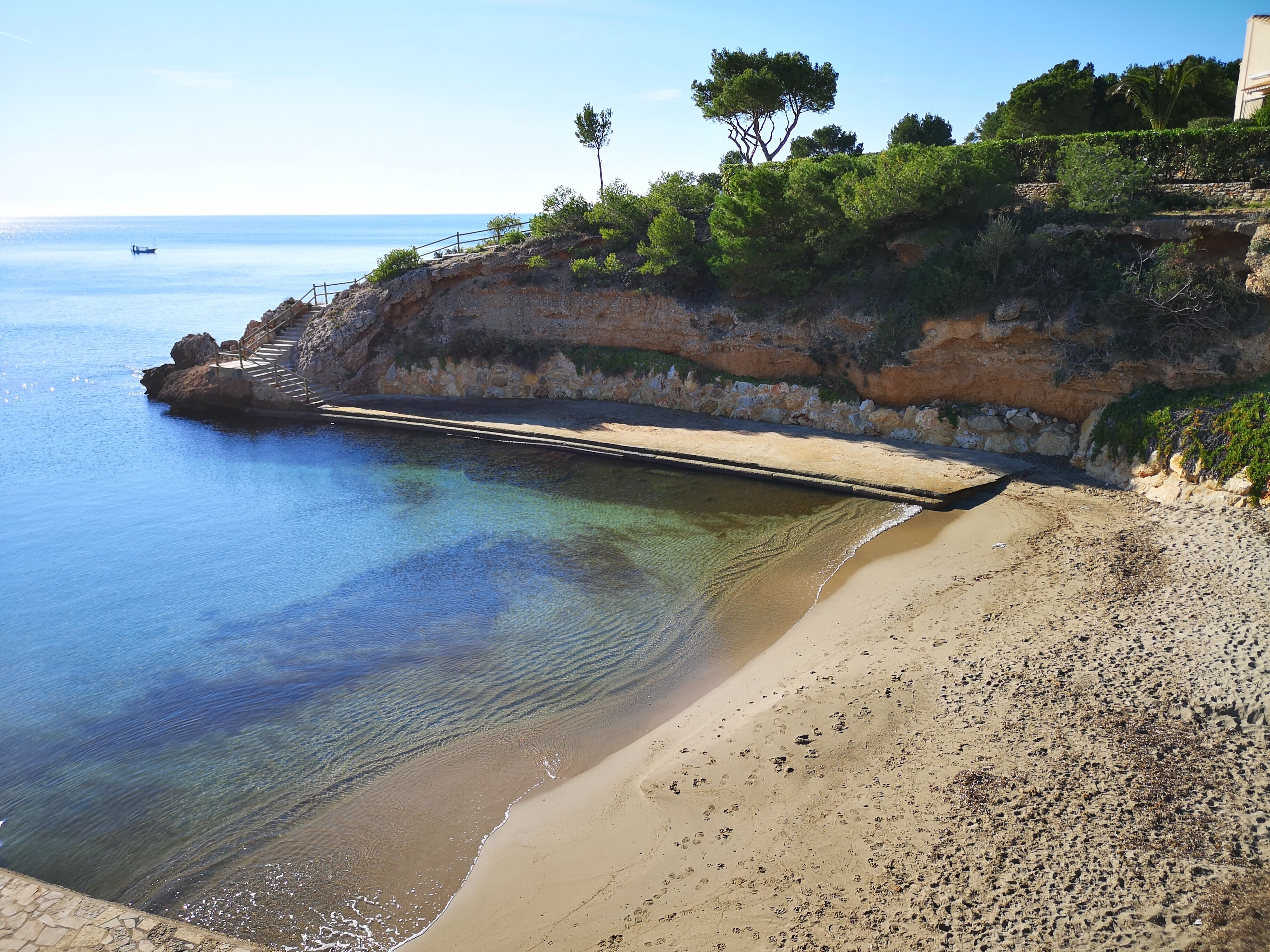 Ein Strand mit klarem Wasser, einer Treppe, die zum Ufer führt, und einem felsigen Felsen mit Vegetation.