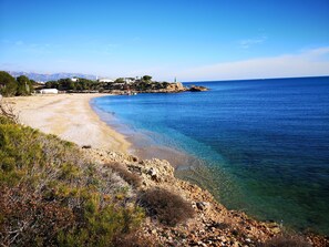 A coastal landscape with a clear beach, rocky shoreline, and a distant mountain range.