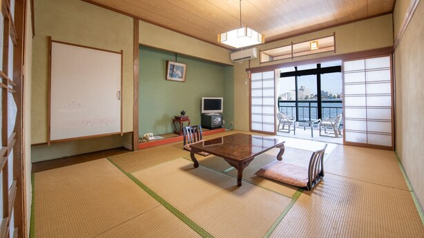 A traditional Japanese room with tatami flooring, a low table, a TV, and a view of a cityscape through large windows. - Hotel Shiiya (Sado)