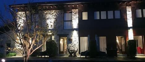 Una casa de dos pisos con fachada de piedra, un patio con mobiliario exterior y una entrada bien iluminada.