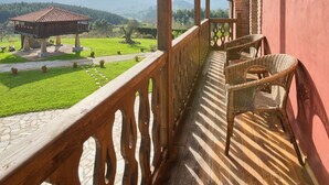 Un balcon doté d’une balustrade en bois, de chaises en osier et offrant une vue sur un paysage verdoyant et des montagnes lointaines.