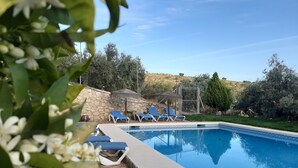 Piscina con sillas reclinables azules, pared de piedra y estructura con techo de paja.