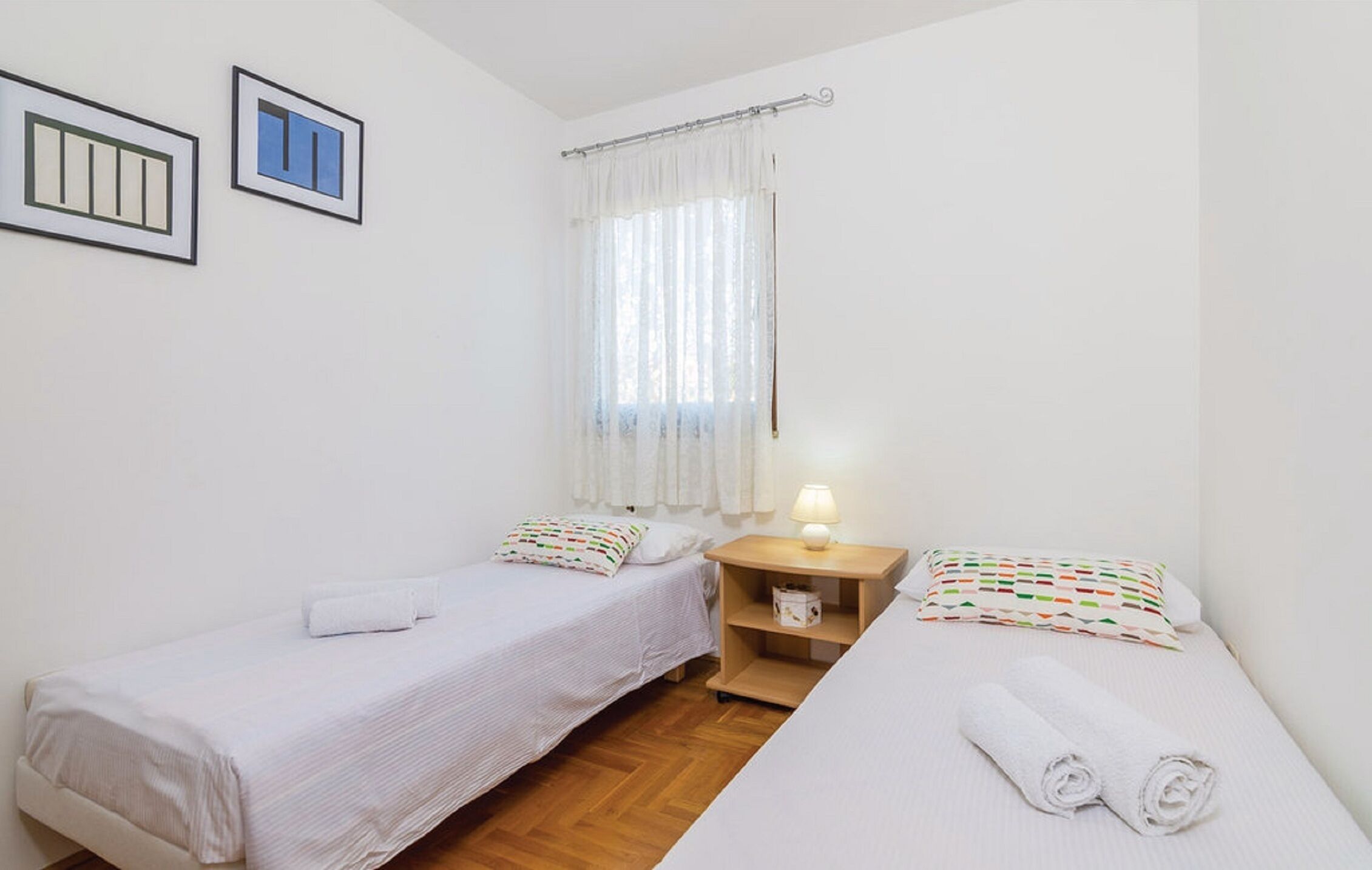 Una habitación pequeña y limpia con dos camas individuales, una mesita de noche con lámpara, una ventana con cortinas translúcidas y dos cuadros enmarcados en la pared.