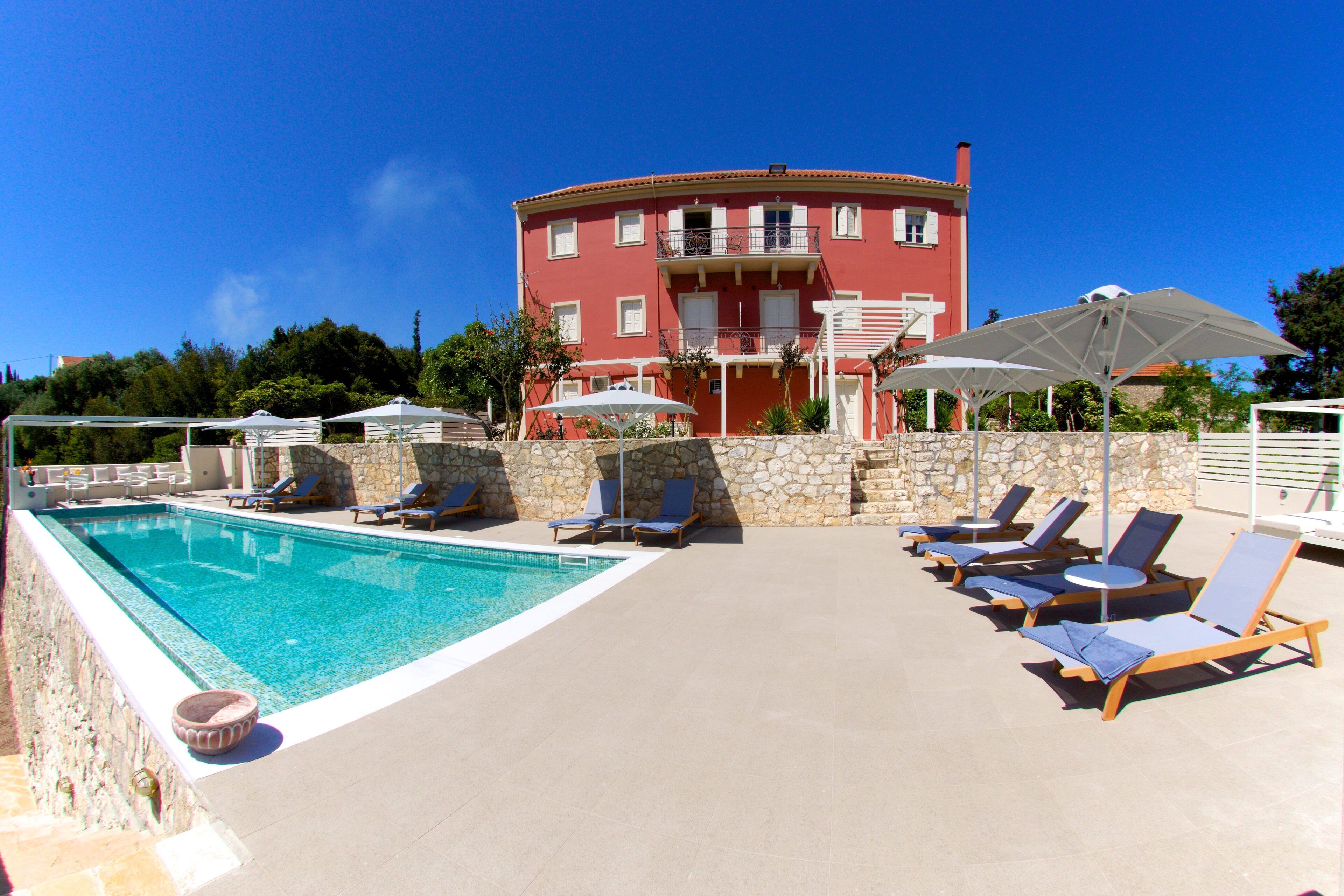 Une piscine avec des chaises longues et des parasols, un mur de pierre et, au fond, un bâtiment rouge doté de balcons.