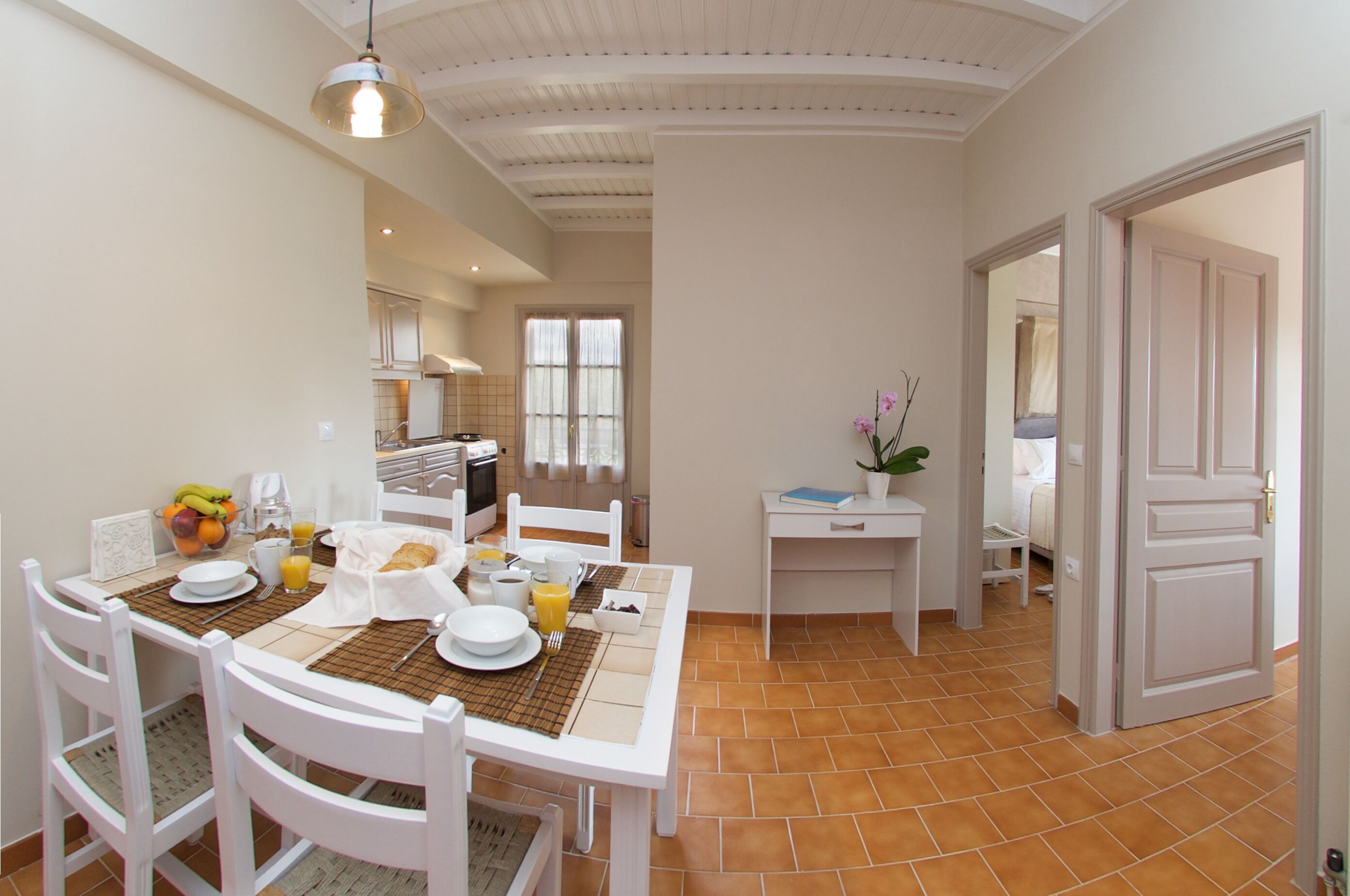 Un coin repas avec une table blanche dressée pour le petit-déjeuner, comprenant des assiettes, des tasses et un panier de fruits.