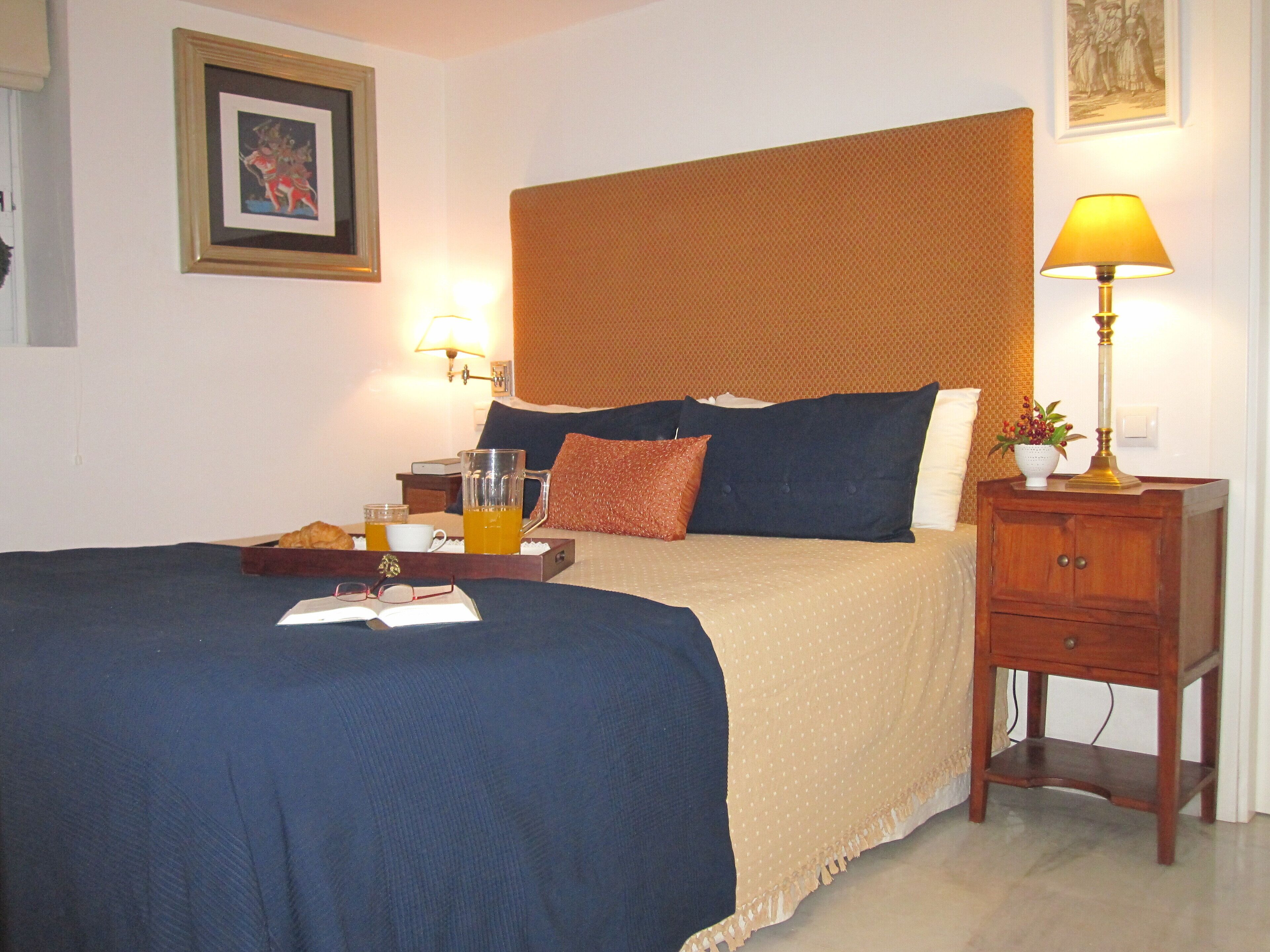 A neatly made bed with a blue bedspread, a wooden nightstand with a lamp, and a framed picture on the wall.