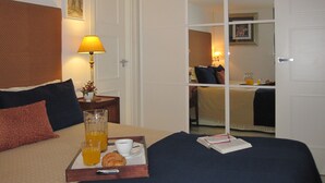 Habitación de hotel con cama, mesita de noche con lámpara, bandeja con productos de desayuno y vista a otra habitación a través de una puerta de vidrio.
