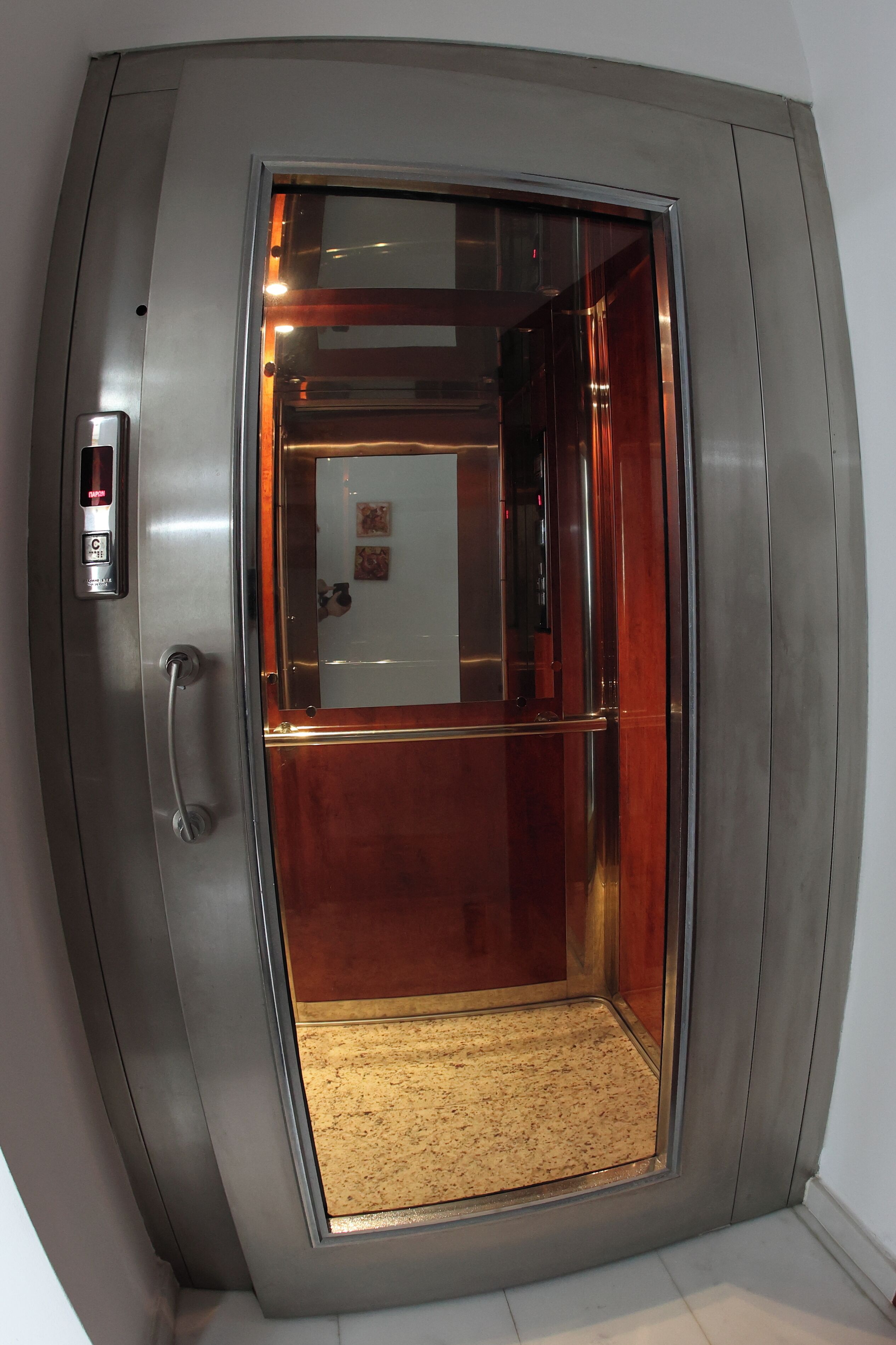 Un ascensor modern amb parets metàl·liques i una porta de vidre, amb un panell de control a l'esquerra.