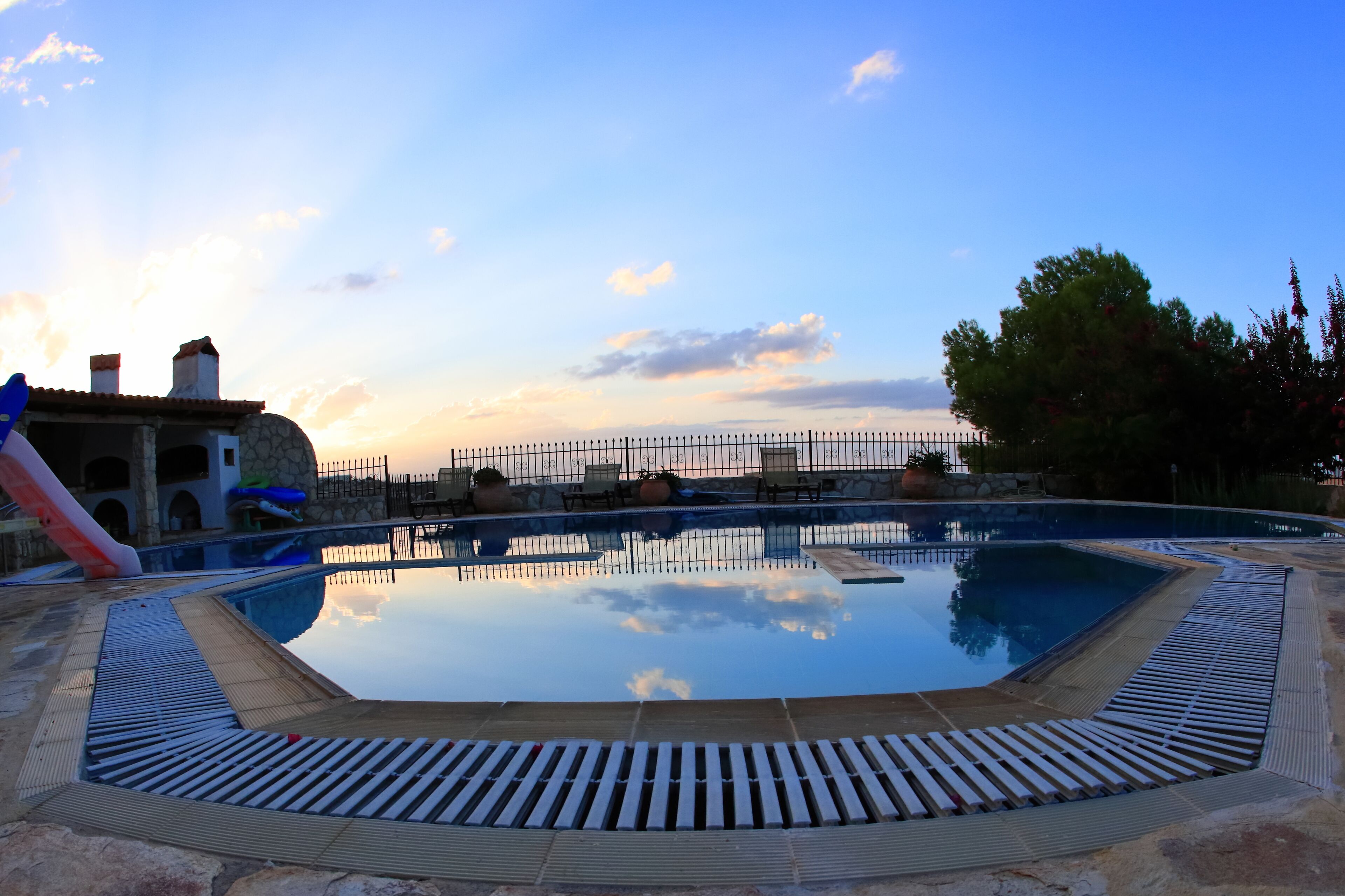 A swimming pool with a surrounding deck, a slide, and a stone building in the background.