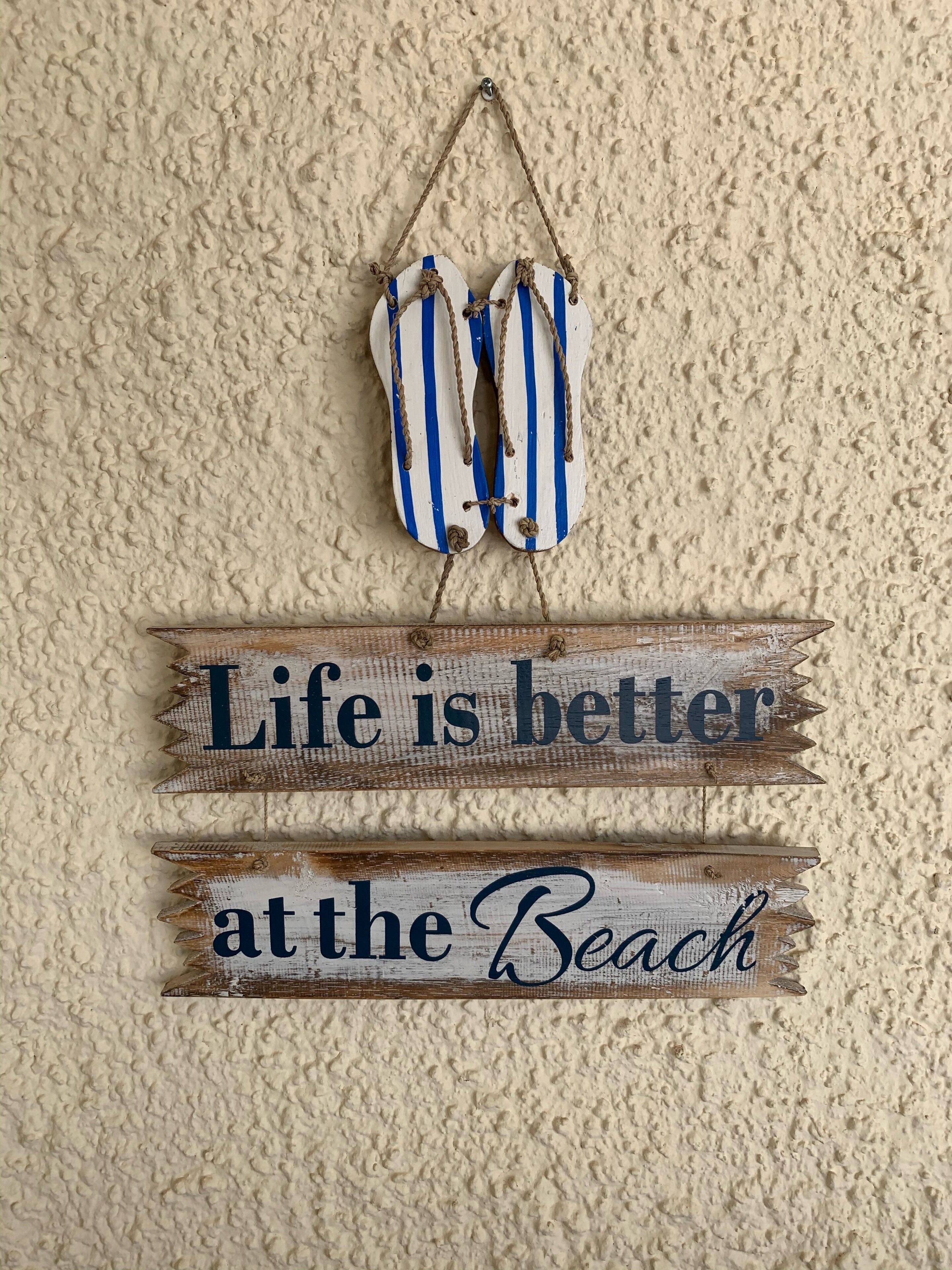 Uma placa de madeira com o texto 'Life is better at the Beach' pendurada numa parede texturizada.