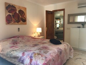 A hotel room with a floral bedspread, a bedside lamp, a wooden door leading to a bathroom, and a white wardrobe.
