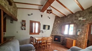 A cozy living room with a stone fireplace, wooden beams, a dining table with chairs, and a television mounted on the wall.