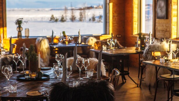 A cozy dining area with wooden tables set for a meal, a large window offering a view of a snowy landscape, and rustic decor including candles and a fur throw. - Torfhús Retreat (Bláskógabyggd)