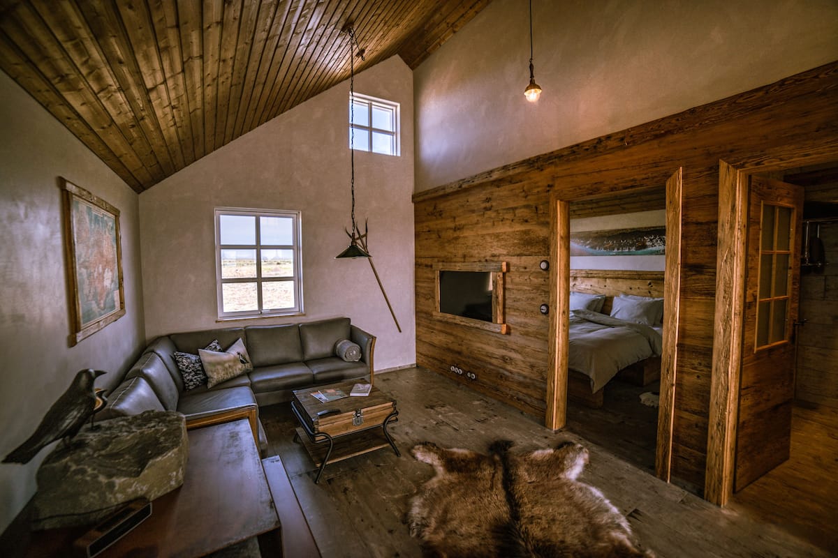 A rustic cabin interior with a wooden ceiling, a sofa, a bed, and a dining table.