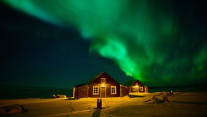 A wooden cabin with a lighted entrance, surrounded by snow, under a night sky with green aurora. - Torfhús Retreat (Bláskógabyggd)