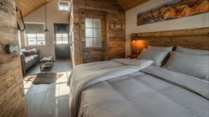 A rustic bedroom with a large bed, wooden walls, a painting, and a view of a room with a sofa and a window. - Torfhús Retreat (Bláskógabyggd)