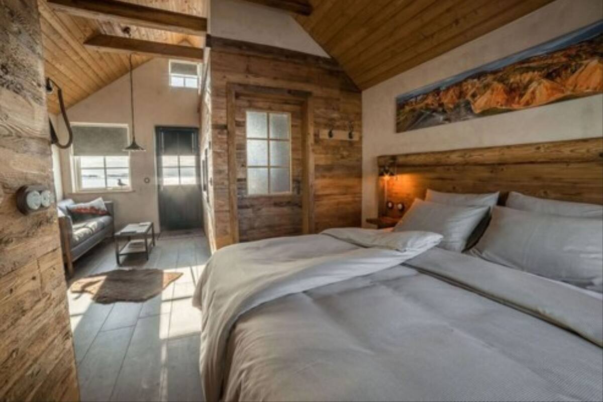A rustic bedroom with a large bed, wooden walls, a painting, and a view of a room with a sofa and a window.