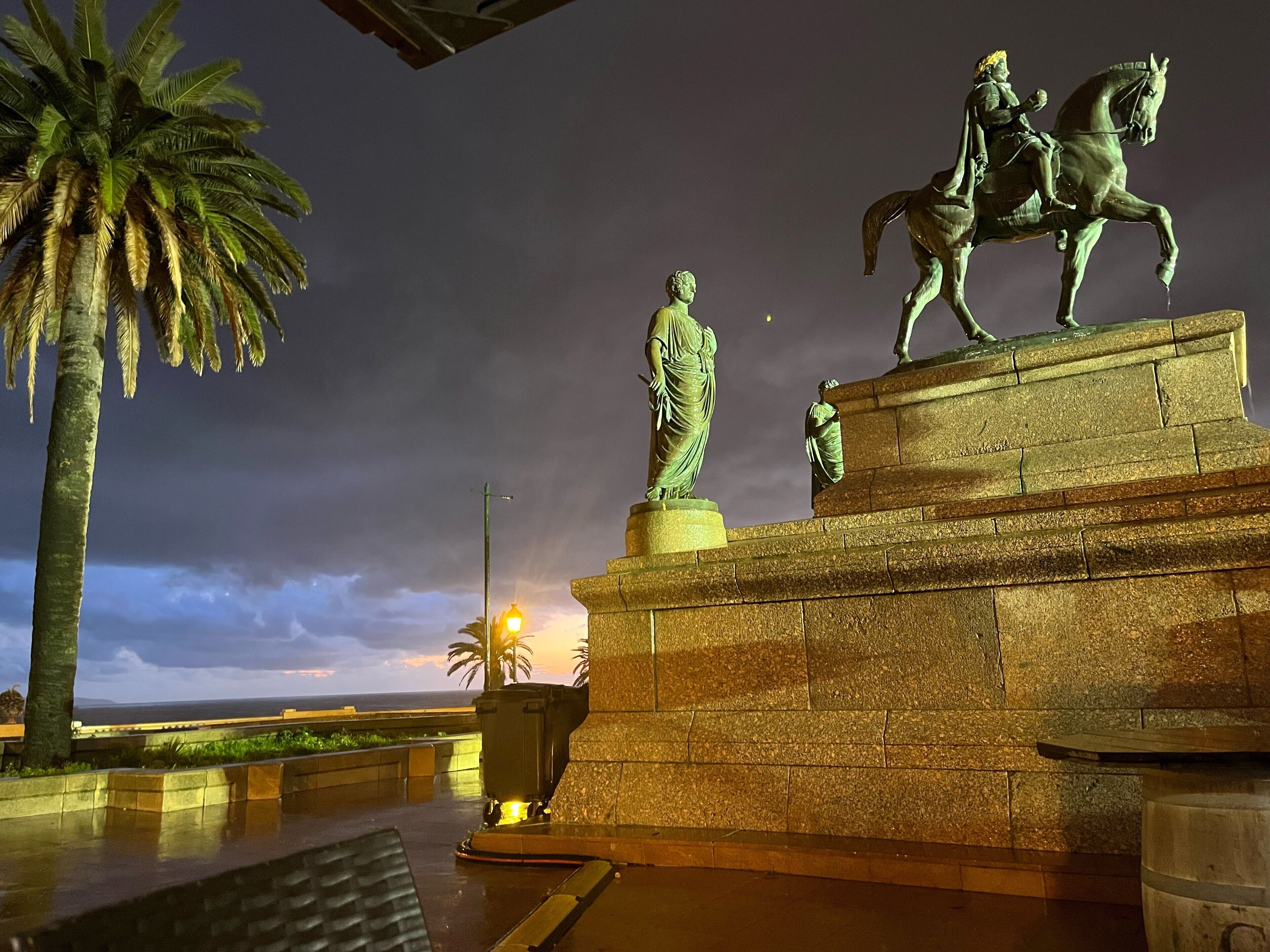 A nighttime view of a plaza with a statue of a man on horseback and another statue of a woman.
