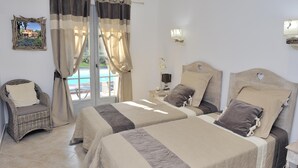 A bedroom with two beds, a wicker chair, a nightstand with a lamp, and a view of the outdoors. - House A Sùvara ***** private pool, park with trees 15 hectares, 5 min PORTICCIO (Cauro)