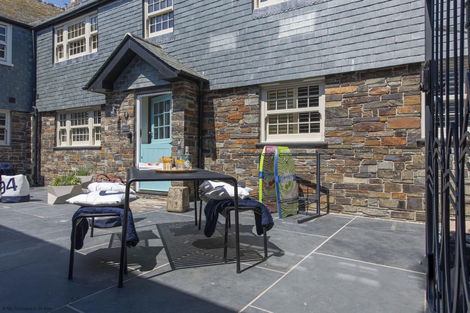 A stone building with a blue door and windows, outdoor seating with chairs and a table, and a surfboard rack.