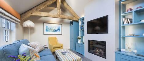 A cozy living room with a blue sofa, a yellow armchair, and a striped ottoman. There is a fireplace, a bookshelf, and a framed picture on the wall.
