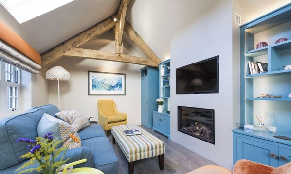 Ein gemütliches Wohnzimmer mit einem blauen Sofa, einem gelben Sessel und einem gestreiften Hocker. Ein Kamin, ein Bücherregal und ein gerahmtes Bild sind an der Wand zu sehen.