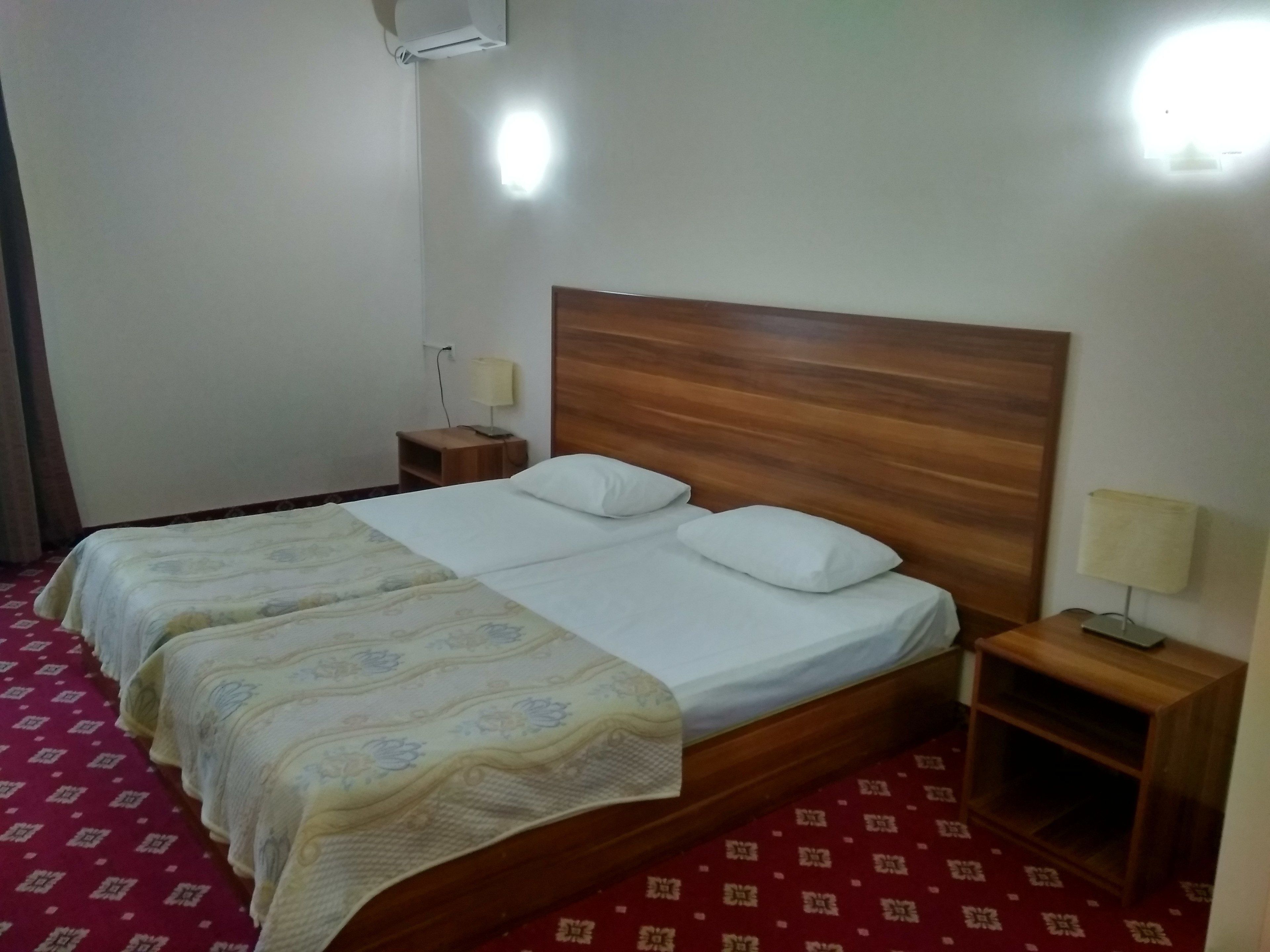 Une chambre d’hôtel avec une tête de lit en bois, un lit simple avec une literie blanche, une table de chevet avec une lampe et un tapis rouge à motifs.