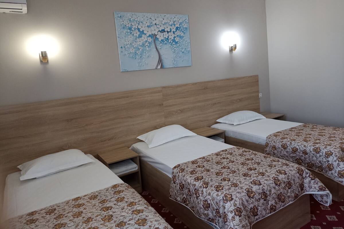 A hotel room with three single beds, a wooden headboard, a floral-patterned bedspread, a wall-mounted painting of a tree, and two wall-mounted lights.