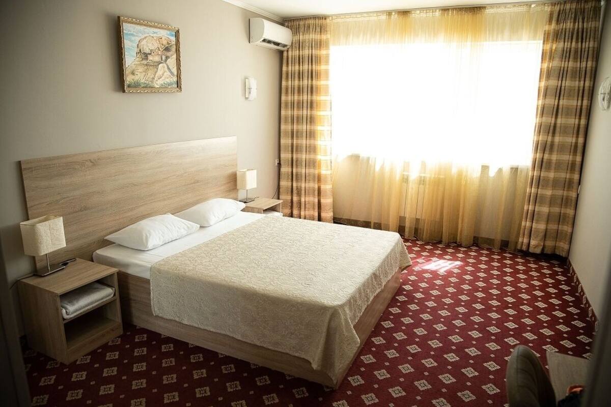 A hotel room with a wooden headboard, a large bed with white linens, a bedside table with a lamp, and a window with curtains.