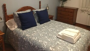 Una cama bien hecha con una colcha estampada, dos almohadas, un aparador, un espejo y un jarrón con flores.