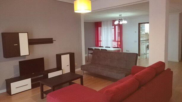 A modern living room with a grey sofa, red armchairs, a coffee table, and a TV on a black stand. There is a dining area in the background with a table and chairs. - Apartment for groups in Logroño (Logroño)