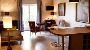 Ein gemütliches Wohnzimmer mit einer Couch, einem Esstisch und einem Sessel. Ein Fenster mit Vorhängen, eine Lampe und ein Bild an der Wand sind zu sehen.