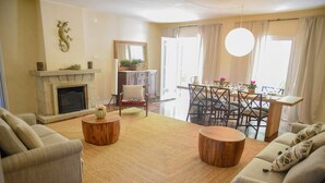 A living room with a fireplace, sofas, a dining area with a table and chairs, and a large window with curtains.
