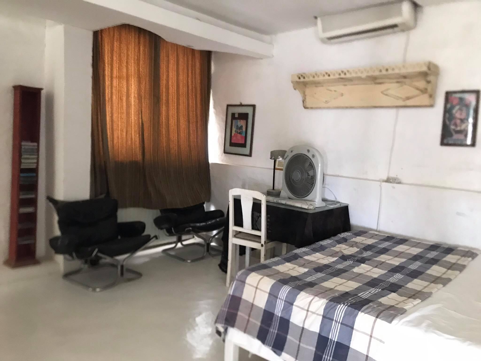 Habitación de hotel con una cama, un escritorio con ventilador, una silla, una estantería y una ventana con cortinas.