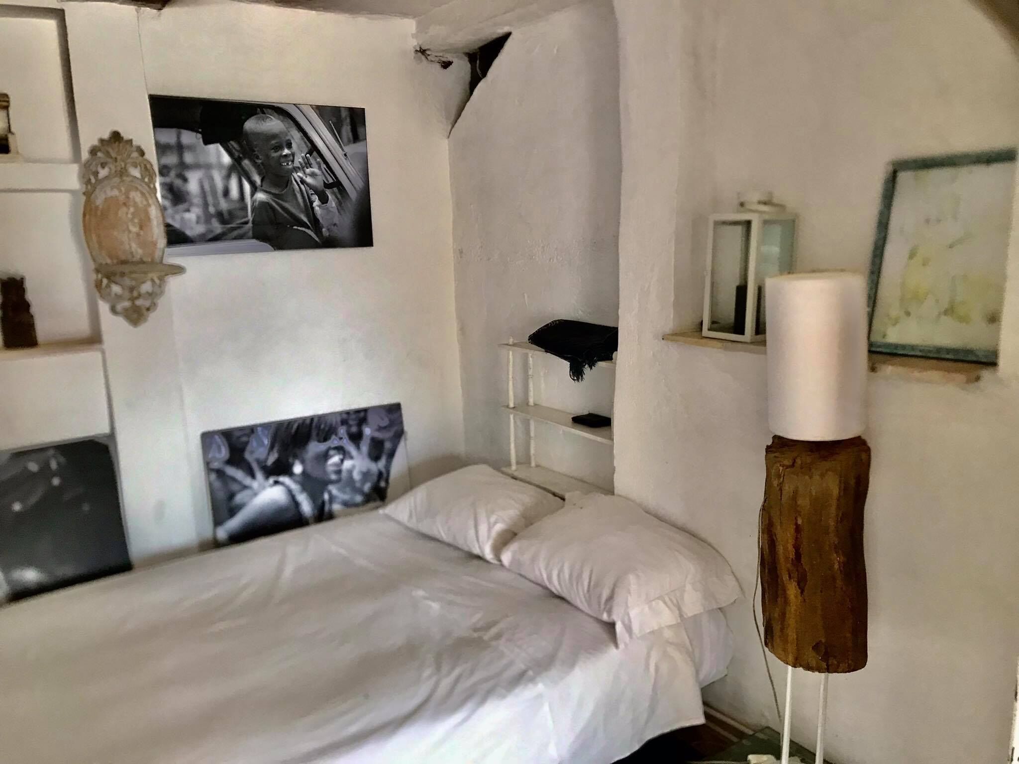 A bedroom with a bed, two black-and-white photographs on the wall, a white lamp, and a shelf with a small item.