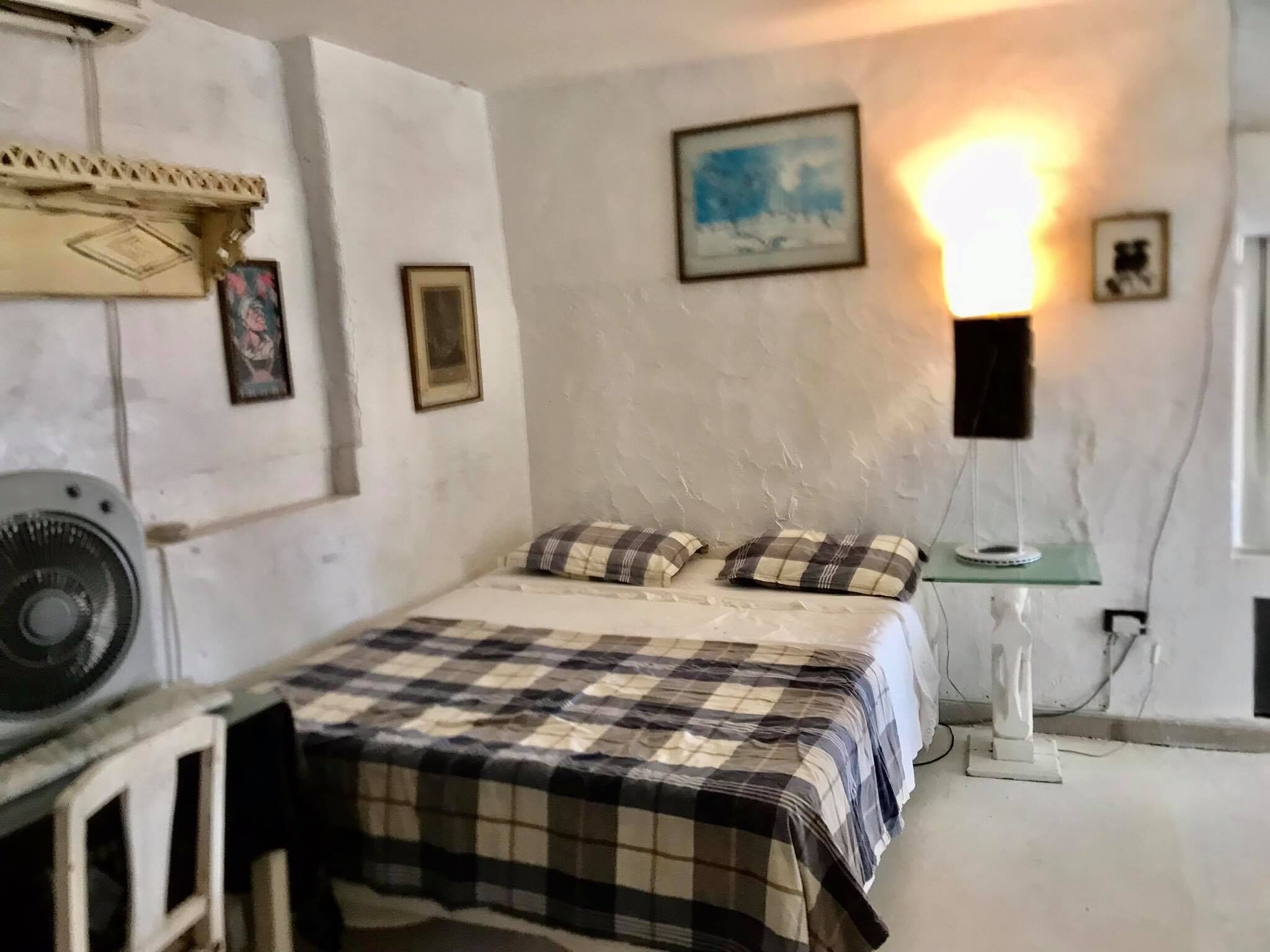 Ein Schlafzimmer mit einem Bett, einem Standventilator, einer Wandleuchte und einem kleinen Tisch.